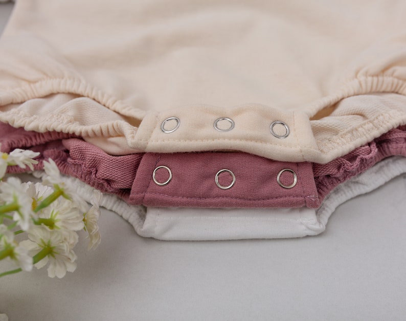 May include: Three baby onesies stacked on top of each other. The top onesie is cream colored, the middle onesie is a dusty rose color, and the bottom onesie is white. All three onesies have silver snaps.