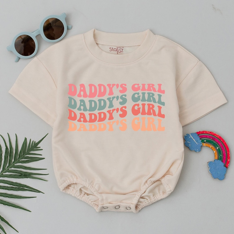 Op de afbeelding: Een cr&egrave;mekleurige romper met korte mouwen met de tekst "Daddy's Girl" die vier keer wordt herhaald in een retro-lettertype. De tekst is in roze en groene tinten.