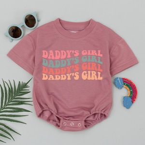 Op de afbeelding: Roze baby romper met de tekst "Daddy's Girl" die vier keer wordt herhaald in een retro lettertype met verschillende kleuren letters.