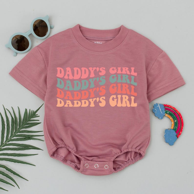 Op de afbeelding: Roze baby romper met de tekst "Daddy's Girl" die vier keer wordt herhaald in een retro lettertype met verschillende kleuren letters.