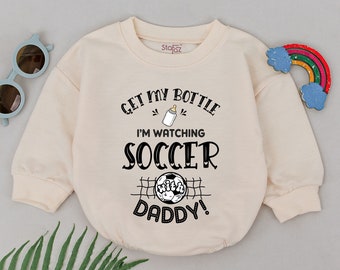 Puede incluir: Un body de bebé de color crema con el texto "Get my bottle I'm watching soccer with daddy!" en negro. El body tiene un gráfico de un biberón, un balón de fútbol y una portería de fútbol.