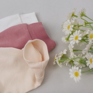 May include: Three folded fabric neck warmers in white, pink, and beige. The neck warmers are made of a soft, ribbed material.