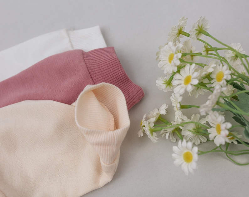 May include: Three folded fabric neck warmers in white, pink, and beige. The neck warmers are made of a soft, ribbed material.