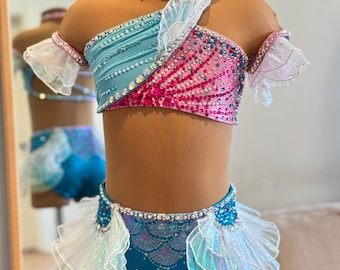 May include: A mermaid-themed dance costume in shades of teal, pink, and white. The top is asymmetrical with a ruffled sleeve and embellished with sequins and rhinestones. The matching shorts feature a scale pattern and layered ruffles. A choker with a seashell and starfish completes the look.