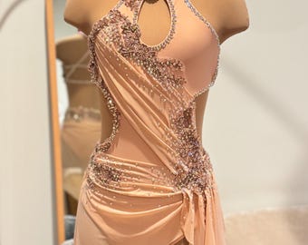 Puede incluir: Un traje de baile de color melocotón con escote halter y un recorte de ojo de cerradura. El traje está adornado con diamantes de imitación y lentejuelas.