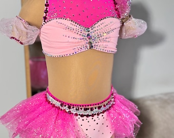 May include: A vibrant pink dance costume with a high neckline and a tutu skirt. The top is embellished with rhinestones and features sheer, ruffled sleeves. The tutu is layered with glittery tulle. The costume is displayed on a mannequin.