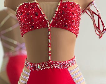Puede incluir: Un traje de baile rojo con un top corto y bragas de talle alto adornados con cristales, unidos por una tira vertical. La prenda incluye aberturas laterales de malla color carne con filas horizontales de cristales y un accesorio para el brazo.