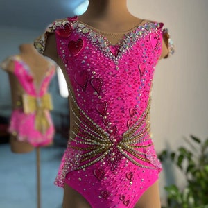Könnte beinhalten: Ein rosa Trikot mit Herz-Design und goldenen Akzenten. Das Trikot ist mit Strasssteinen besetzt und hat eine Mesh-Einstellung im Rücken.