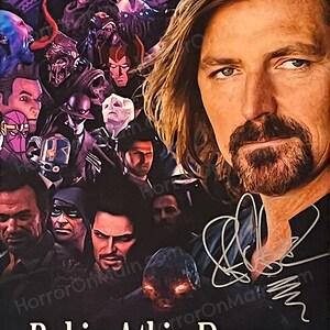 May include: A poster featuring a portrait of Robin Atkin Downes, with a collage of character images in the background. The poster has a dark background with the name "Robin Atkin Downes" in white text at the bottom, and a signature.