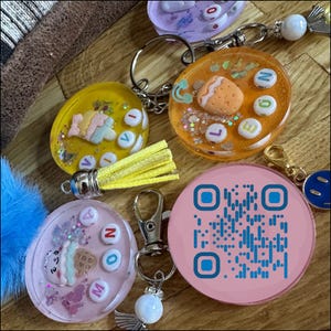 Könnte beinhalten: Eine Sammlung bunter Schlüsselanhänger aus Kunstharz mit verschiedenen Designs. Einer ist gelb mit einer Quaste, ein anderer orange mit einer Erdbeere und ein dritter rosa mit einem QR-Code. Jeder Schlüsselanhänger hat einen silbernen Verschluss.