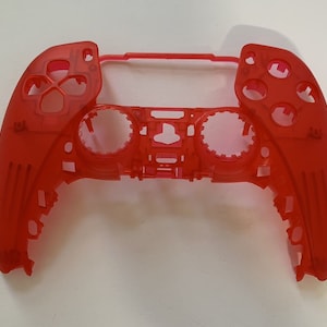 May include: A red plastic shell for a video game controller. The shell is translucent and has a textured surface. The shell is missing the buttons and joysticks.