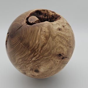 Puede incluir: Objeto decorativo esférico de madera con un acabado marrón claro natural. La veta de la madera es visible y hay una gran abertura en la parte superior. El objeto tiene una estética rústica y orgánica.