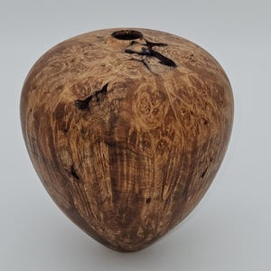 Puede incluir: Un jarrón de madera hecho a mano con forma redondeada y una pequeña abertura en la parte superior. El jarrón está hecho de madera de color marrón claro con un patrón de vetas natural y nudos oscuros, mostrando la textura única de la madera.