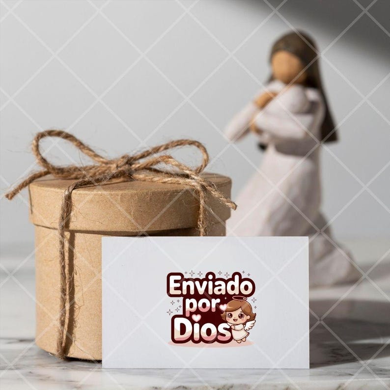 May include: A white card with a brown and pink cartoon angel with the text "Enviado por Dios" on a white background. The card is sitting on top of a brown gift box with twine.