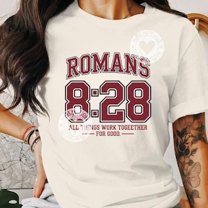 Puede incluir: Camiseta color crema con el texto "ROMANS 8:28" en granate y negro. La frase "ALL THINGS WORK TOGETHER - FOR GOOD" está impresa debajo. La camiseta tiene mangas cortas y un corte relajado.