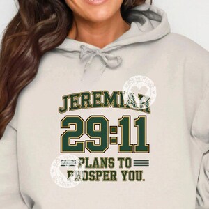 Może przedstawiać: Jasnobeżowa bluza z kapturem z napisem "JEREMIAH 29:11 PLANS TO PROSPER YOU" w zielonych i złotych literach. Bluza ma sznurek i przednią kieszeń.