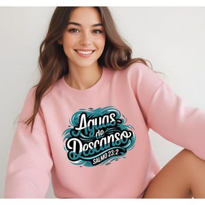 May include: Pink sweatshirt with a teal blue graphic design that says "Aguas de Descanso" with the text "Salmo 23:2" underneath.