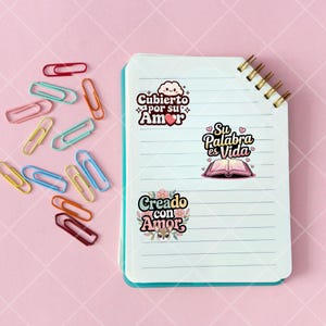 May include: A white notebook with blue lines and a gold spiral binding. Three colorful stickers are on the notebook. The stickers have Spanish text that reads "Cuberto por su Amor", "Su Palabra es Vida", and "Creado con Amor".