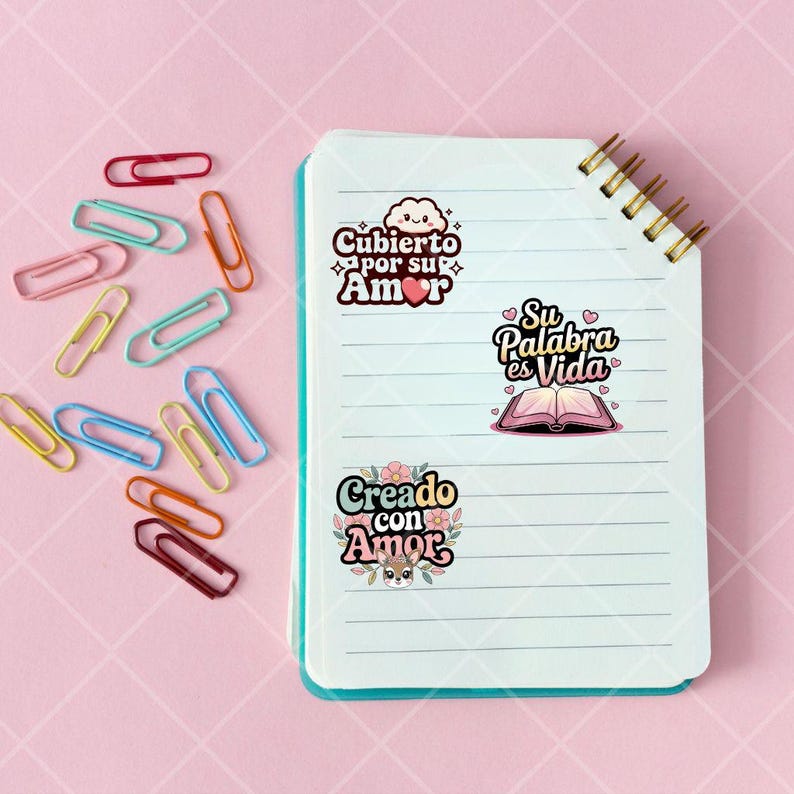 May include: A white notebook with blue lines and a gold spiral binding. Three colorful stickers are on the notebook. The stickers have Spanish text that reads "Cuberto por su Amor", "Su Palabra es Vida", and "Creado con Amor".