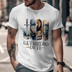 May include: White t-shirt featuring a watercolor design with a sword, open book, lion, and shield. The text "GUERRERO DE FE" is printed below the images. The shirt is made of a soft material.