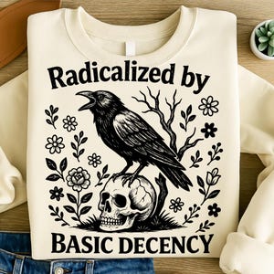 Op de afbeelding: Crèmekleurige sweatshirt met een zwarte afbeelding van een raaf op een schedel, omringd door bloemen en takken. De tekst "Radicalized by BASIC DECENCY" staat boven en onder de ravenillustratie.