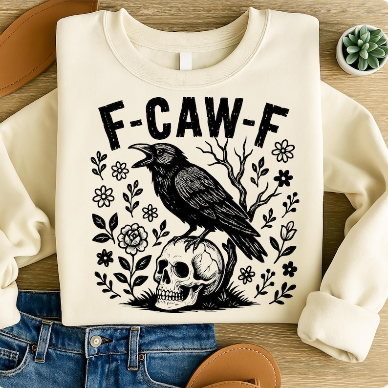May include: Cream-colored sweatshirt with a black graphic of a crow perched on a skull, surrounded by floral and botanical elements. The text "F-CAW-F" is printed above the crow. The sweatshirt is folded on a wooden surface.