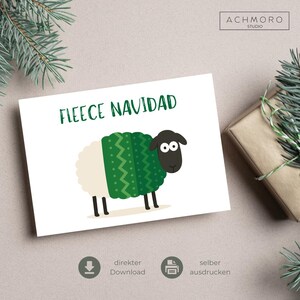 Könnte beinhalten: Eine weiße Karte mit dem grünen Text „FLEECE NAVIDAD“. Sie zeigt ein Cartoon-Schaf mit einem grünen Pullover und einem schwarzen Gesicht. Die Karte liegt auf einer neutralen Oberfläche, mit einem Geschenk und Tannenzweigen.
