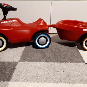 Könnte beinhalten: Rotes Big Bobby-Car Spielzeug mit Anhänger. Das Auto hat ein Lenkrad, einen Sitz und vier Räder mit weißen und gelben Streifen. Der Anhänger ist ebenfalls rot und hat vier Räder mit weißen und orangen Streifen.