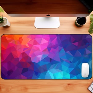 May include: A colorful geometric design mouse pad with a white computer mouse on top of it. The mouse pad is on a wooden desk with a computer monitor, a cup of coffee, and two potted plants.