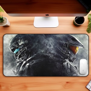 May include: A rectangular mousepad featuring a detailed graphic of two futuristic soldiers in helmets, one with blue visor, the other with an orange visor. The design is predominantly gray and black, with a city silhouette in the background. A white mouse and keyboard are visible.