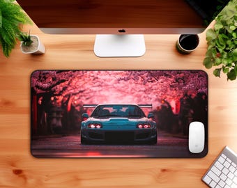 Puede incluir: Alfombrilla de ratón rectangular con un coche deportivo turquesa en una carretera bordeada de cerezos en flor. Los faros del coche están encendidos, sobre un fondo rosa. También se ven un ratón y un teclado blancos.