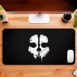 May include: Black gaming mouse pad with a white skull graphic. The skull has a cracked and dripping effect. The mouse pad is on a wooden desk with a computer monitor, a coffee mug, and two potted plants.