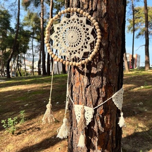 Puede incluir: Un atrapasueños de crochet blanco con cuentas de madera colgando de un árbol. El atrapasueños tiene una borla blanca y una guirnalda de macramé blanca y marrón.