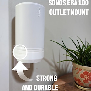 May include: A white Sonos Era 100 outlet mount is shown, mounted on a wall. The mount is designed to hold the speaker securely. The text "SONOS ERA 100 OUTLET MOUNT" and "STRONG AND DURABLE" are visible.