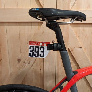 May include: A black bicycle seat with silver rails and a black seat post. The bicycle frame is red and gray. A race number plate with the number 393 is attached to the seat post.