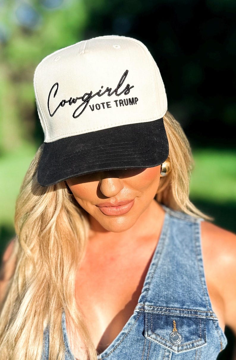 K&ouml;nnte beinhalten: Eine wei&szlig;e und schwarze Baseballkappe mit dem Text "Cowgirls Vote Trump" in schwarzer Stickerei auf der Vorderseite.