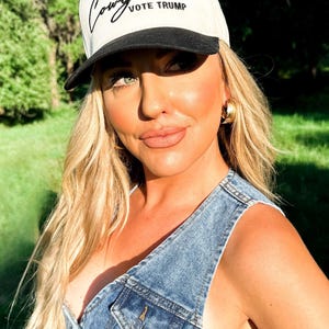 K&ouml;nnte beinhalten: Eine Frau tr&auml;gt eine wei&szlig;e und schwarze Baseballkappe mit dem Schriftzug "Cowgirls Vote Trump" in schwarzer Stickerei. Sie tr&auml;gt eine Jeansweste und hat blonde Haare.