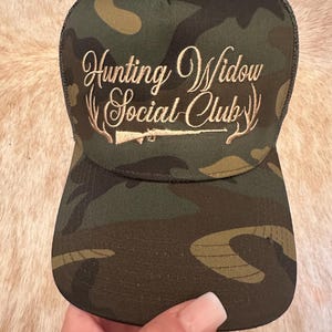 Pode incluir: Um boné de caminhoneiro camuflado com um logotipo bordado em ouro que diz "Hunting Widow Social Club" com um rifle e chifres.