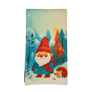 May include: A decorative kitchen towel featuring a whimsical winter scene. A gnome with a red hat and scarf stands in a snowy forest with a hedgehog. The towel has a blue and white color scheme with a waffle weave texture.