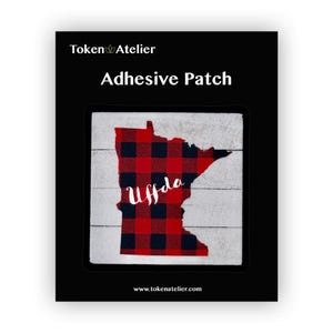 May include: Red and black plaid Minnesota state outline with the word "Uffda" in white lettering.  The patch is on a white wood background.  Adhesive patch.