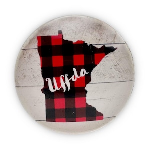 May include: A round ceramic plate with a white background and a red and black plaid outline of the state of Minnesota. The word "Uffda" is written in white script inside the outline.