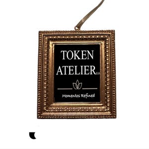 May include: A decorative, rectangular frame with a gold finish. The frame surrounds a black background with the text "TOKEN ATELIER" and "Momentos Refined" in white. A gold ribbon is attached for hanging.