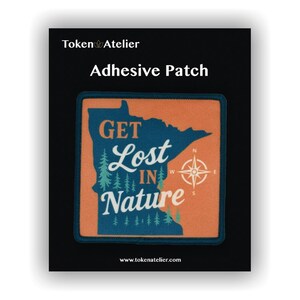 May include: Adhesive patch with a design featuring the text "GET Lost IN Nature" over an orange and blue map outline. A compass rose is on the right. The patch is on a black background with the words "Token Atelier" and "Adhesive Patch".