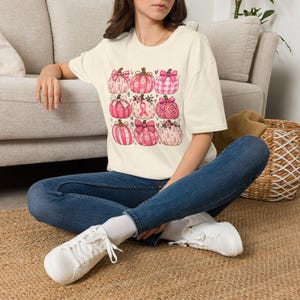 May include: A cream-coloured short-sleeve t-shirt featuring a graphic print of nine pumpkins in various shades of pink. The pumpkins are decorated with bows, floral patterns, leopard print, plaid, and breast cancer awareness ribbons.