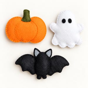 May include: Three felt ornaments featuring a bright orange pumpkin with a dark green stem, a white ghost with two black circular eyes, and a black bat with light gray inner ears. Each piece is constructed from felt with visible stitching.