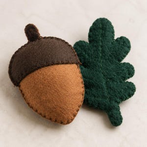 Puede incluir: Una bellota de fieltro y una hoja de roble de fieltro. La bellota presenta un casquillo marrón oscuro y un cuerpo de nuez marrón claro con bordes cosidos visibles. La hoja de roble es de color verde bosque profundo con detalles de nervaduras.