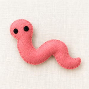 May include: A pink felt worm plush toy featuring a curved, segmented body shape and two small, circular black felt eyes positioned on the head. The edges of the soft, pink material are joined together with visible light-colored stitching.