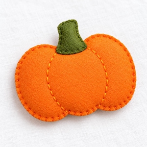 Puede incluir: Un adorno de calabaza de fieltro con un cuerpo de color naranja brillante y costuras naranjas visibles a lo largo de los bordes y segmentos. La calabaza tiene un tallo de color verde oliva oscuro en la parte superior, hecho de fieltro.