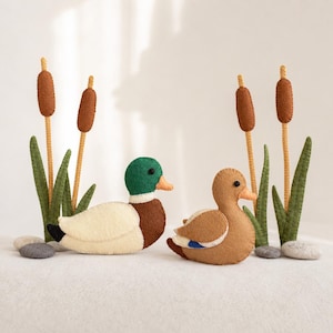 May include: Two felt ducks and four cattail plants made of felt. One duck has a green head, white body, and brown chest. The other duck is light brown with a small blue patch on its wing. Both have orange beaks and black bead eyes.