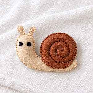 May include: A felt snail ornament featuring a light beige body with two upright antennae and two small black circular eyes. The snail has a prominent, spiraled shell in a deep brown color, with visible stitching along the edges of the felt.